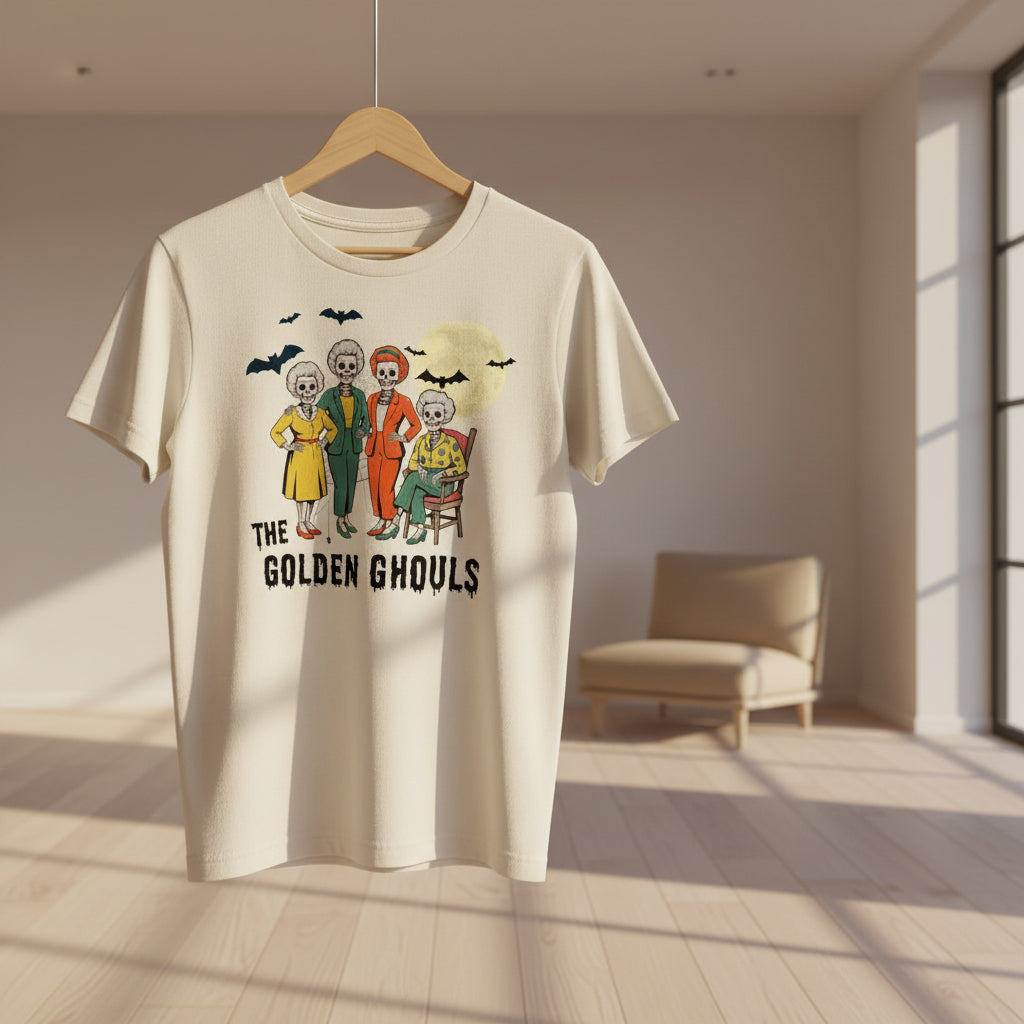 An off-white short-sleeve t-shirt featuring a colorful graphic of 'The Golden Ghouls' characters and bats, displayed against a black background with white spiderwebs.