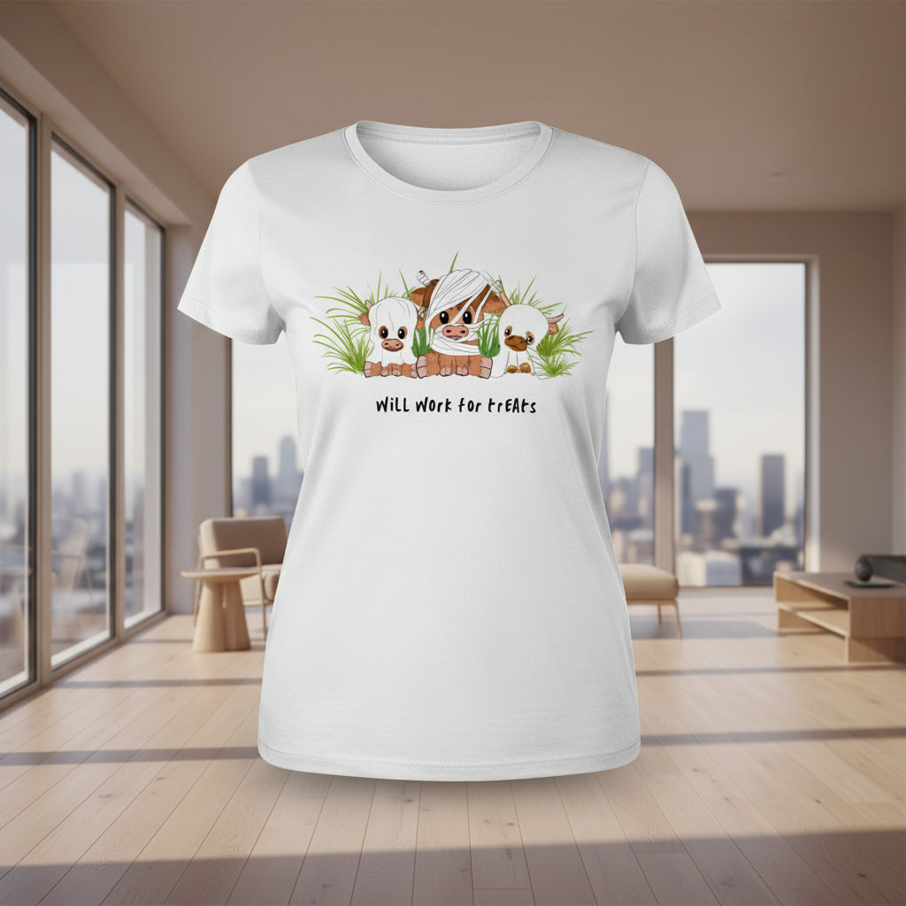 A white short-sleeve t-shirt laid flat on a wooden surface, featuring a cartoon graphic of three piglets in a grassy field with the text 'Will Work for treats'. The shirt is styled with a yellow sunflower, white sneakers, and blue denim jeans.